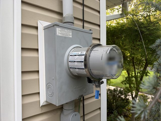GenerLink transfer-switch collar and electric meter installed on the exterior wall of a residence with conduit runs and utility seal tag